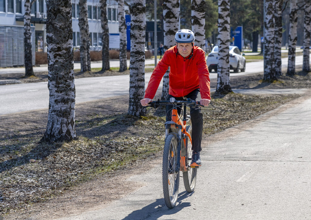 Pyöräilykypärään ja ulkoiluasuun pukeutunut Antti Rovamo pyöräilee oranssilla maastopyörällä koivujen reunustamalla pyörätiellä. Taustalla näkyy OSAOn Kaukovainion kampuksen rakennuksia.