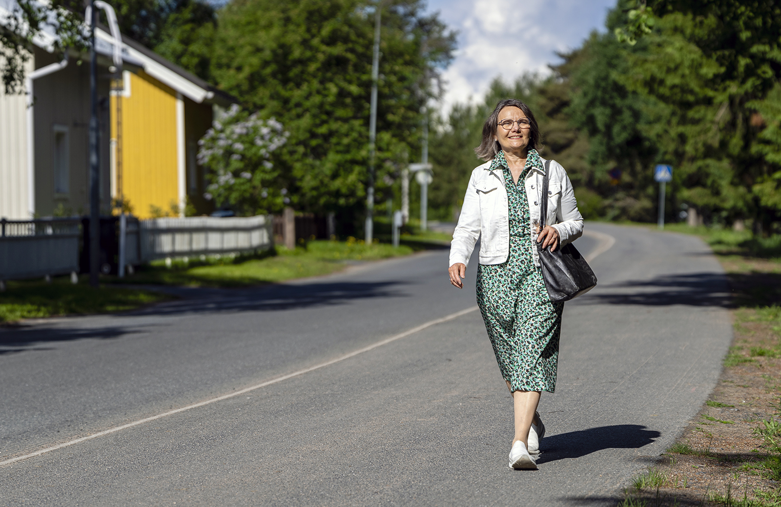 Kirsi Joki-Tokola kuvattuna kävelemässä pyörätiellä päällään valkoinen farkkutakki ja virheä kauluspaita, olallaan musta käsilaukku.